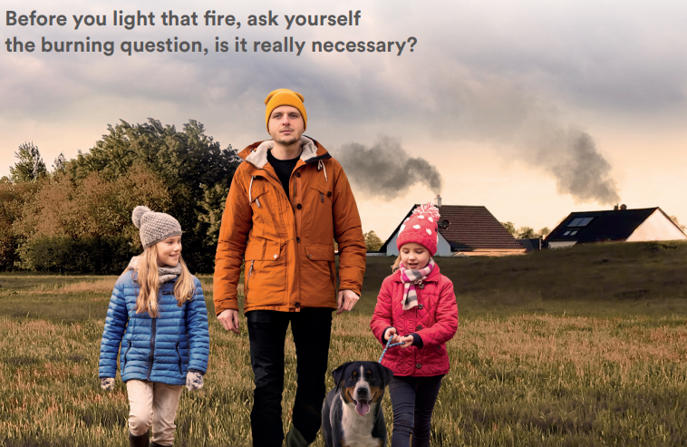 Image shows a father and two daughters walking their dog in a field, dressed for Autumn weather. In the background are two houses with dark smoke rising from their chimneys. The text reads "Before you light that fire, as yourself the burning question, is it really necessary?"