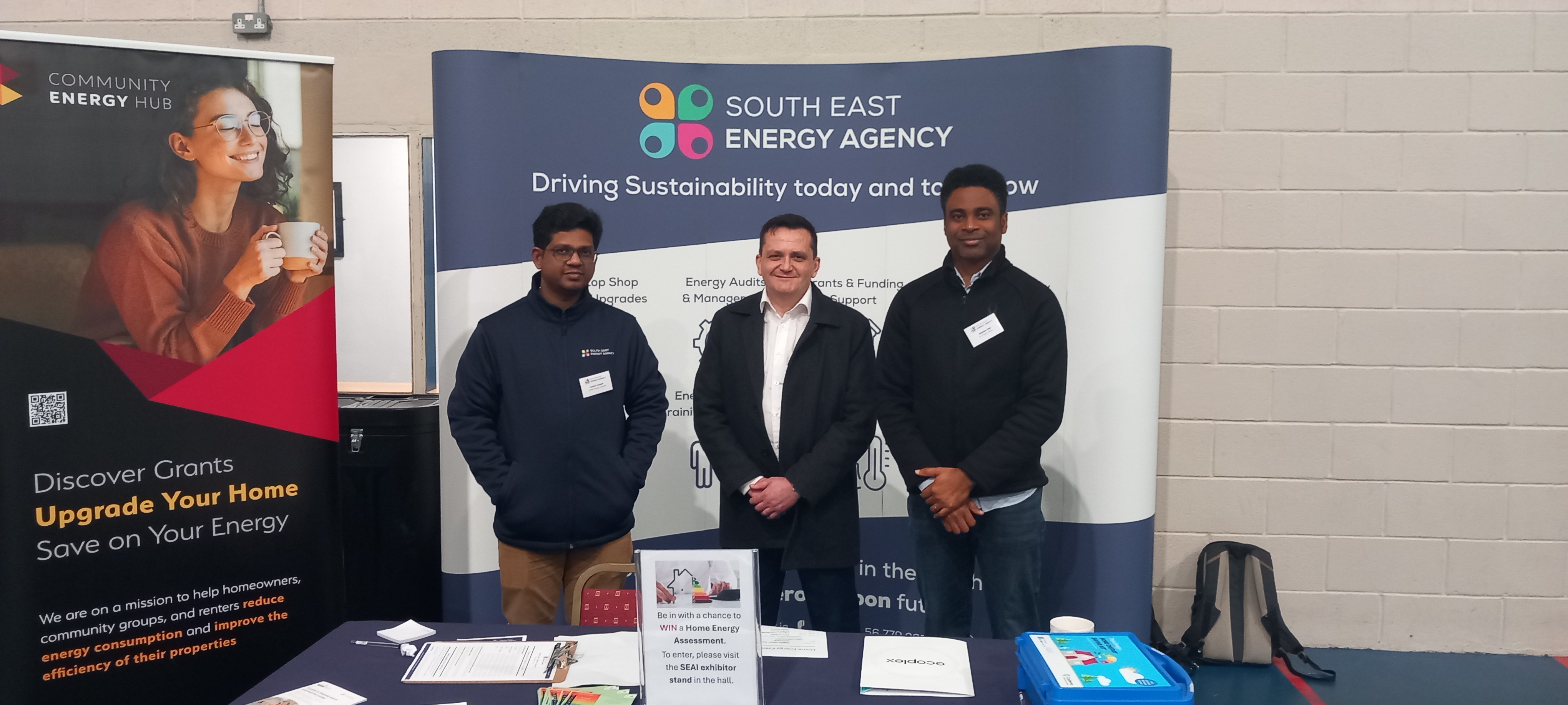 Image shows three men standing and smiling in front of a backdrop sign that reads 