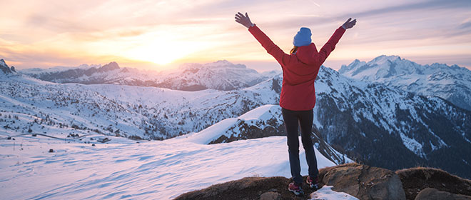 Frau blickt inmitten einer schneebedeckten Berglandschaft dem Sonnenaufgang entgegen