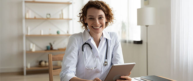 Junge Ärztin sitzt in ihrer Praxis und hält lächelnd ein Tablet in der Hand