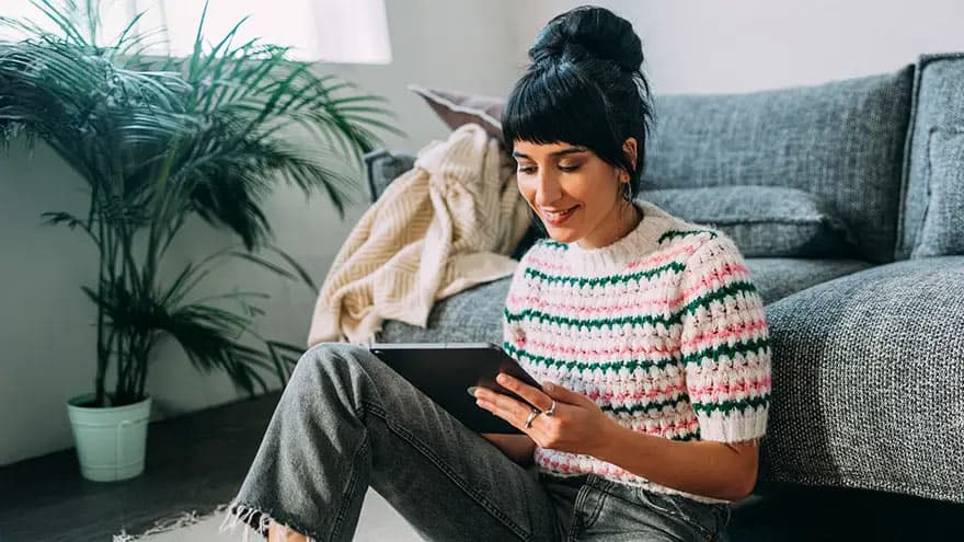 Eine junge Frau in einer Wohnung sitzt mit einem Tablet in der Hand an ein Sofa gelehnt auf dem Boden.