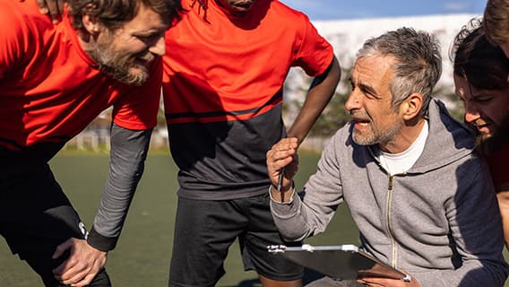 Ein Trainer mit Stift und Klemmbrett spricht auf einem Sportfeld mit seinem Team.