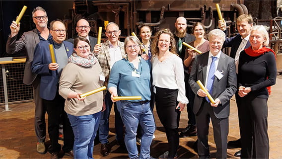 Menschen stehen in einer Gruppe, lächeln freundlich in die Kamera und jede*r hält einen Staffelstab in die Höhe.