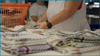 Das Bild zeigt den Oberkörper einer Frau in einer Wäscherei. Die Frau faltet Handtücher zusammen.