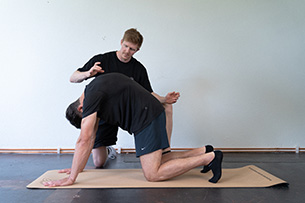 Qualifizierung zum/zur Rücken-Trainer:in Ein Trainer hilft einem Mann, der auf einer Matte sitzt und sich in eine Katzen-Dehnübung befindet, indem er ihm Anweisungen gibt und ihn anleitet.