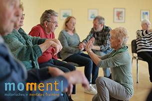 Qualifizierung "moment! - motorisches und mentales Training für Menschen mit Demenz" Ein Gruppe älterer Menschen sitzt im Stuhlkreis eine Übungsleiterin gibt Anweisungen zu Bewegung in der Mitte