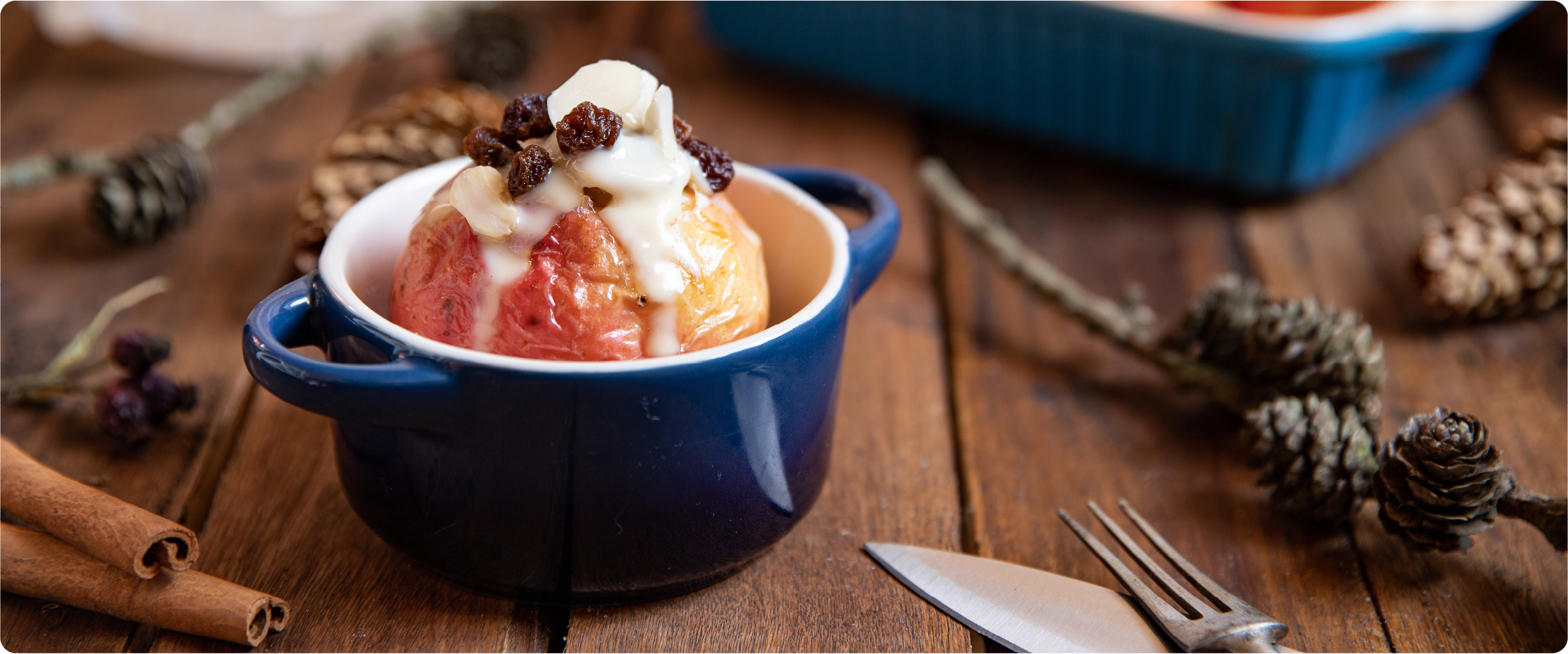 Frisch gebackener Bratapfel in einer kleinen blauen Keramikform, gefüllt mit Rosinen und Mandeln und übergossen mit Vanillesauce; arrangiert auf einem Holztisch mit Zimtstangen und Tannenzapfen für eine winterlich-gemütliche Atmosphäre.