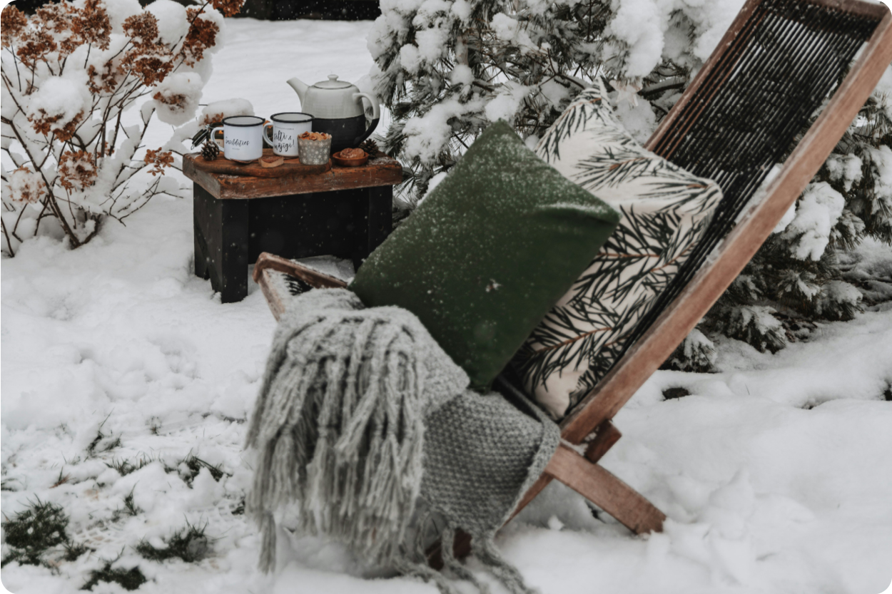 Gemütliche Winterstimmung im Garten mit Liegestuhl, Decken und heißem Tee im Schnee.