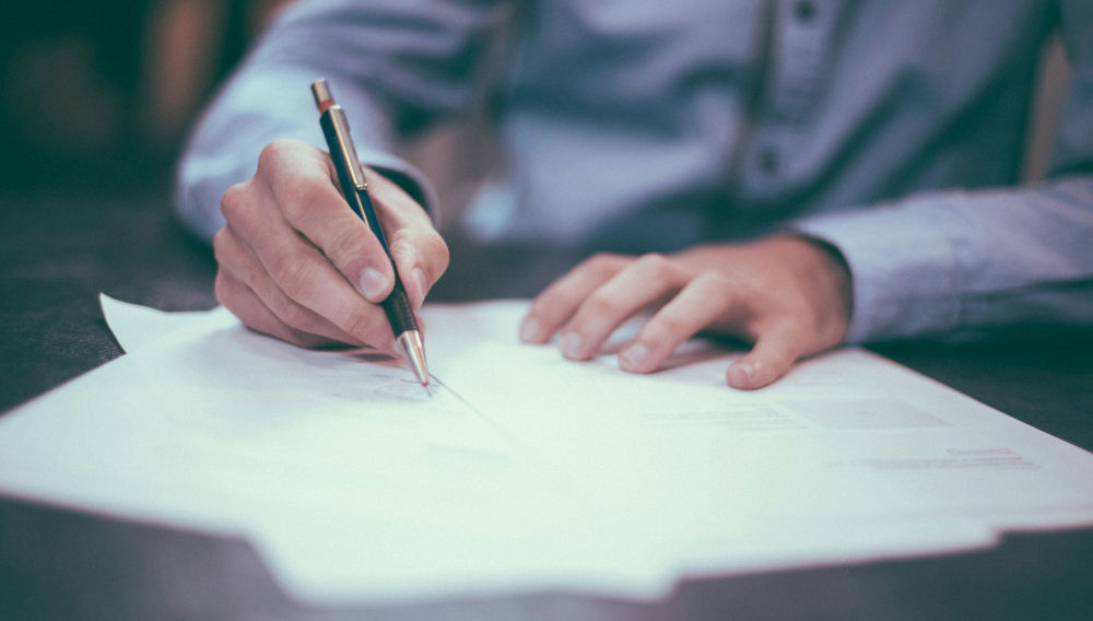A person in business attire is signing a paper; the image detail shown here focuses on hand, pen and paper (Photo: Scott Graham on Unsplash)