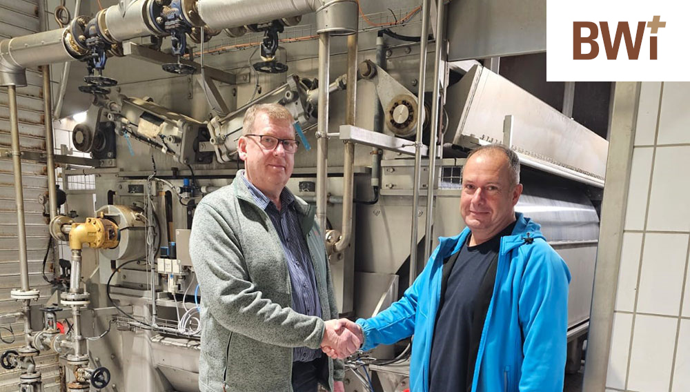 Two men shaking hands in front of pipes in a brewery
