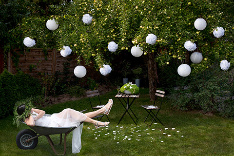 Frau mit einem grünem Blumenkranz im Haar liegt verträumt im weissen Hochzeitskleid in einer Schubkarre im Garten