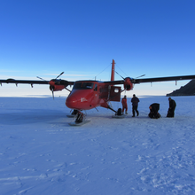 People load equipment onto a red aeroplane that has landed in the middle of the snowy Antarctic tundra.