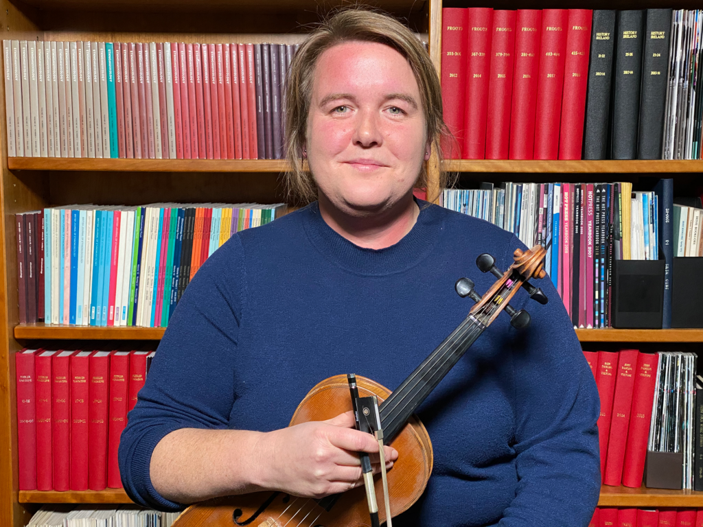 A person posing in front of a book case holding a fiddle in front of them