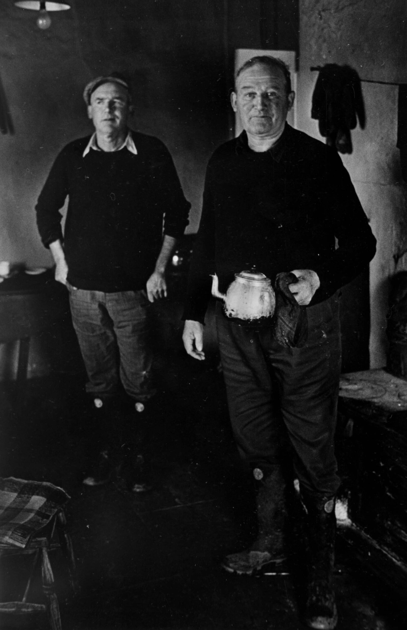 Black and white photograph of two men in dark jumpers standing in a cottage. The man on the right is holding a teapot.