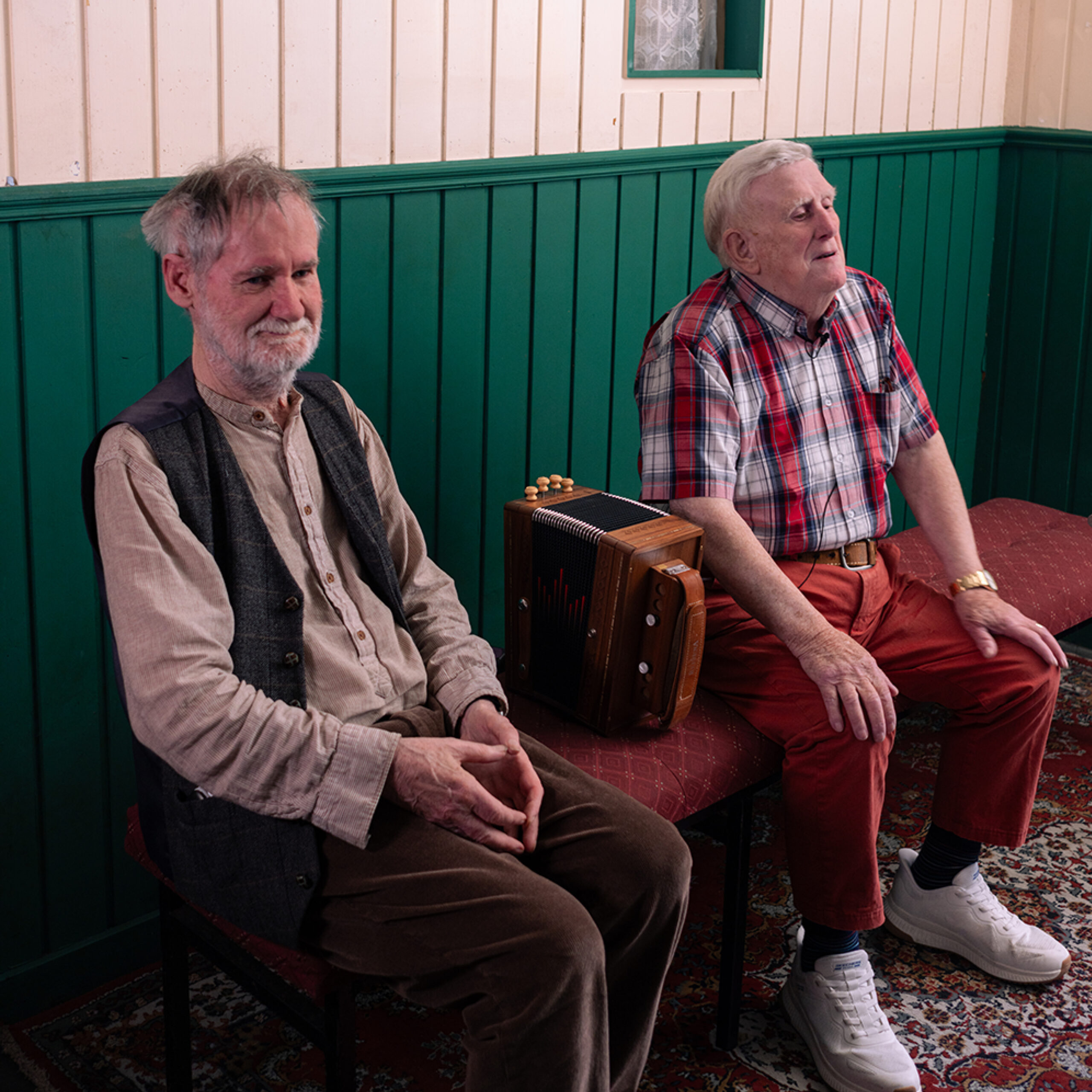 An elderly man in a vest sits on a bench beside another elderly man. An accordion is placed on the bench between the two of them.