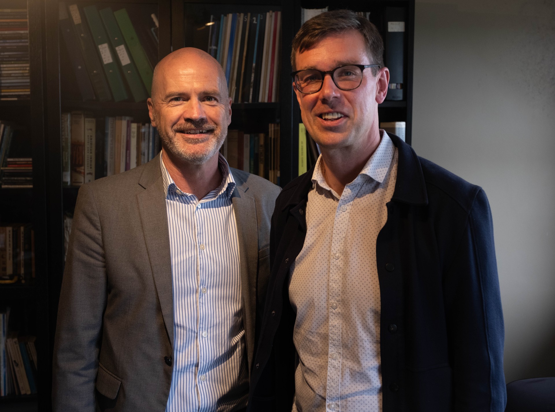 Two men stand in front of a bookshelf.