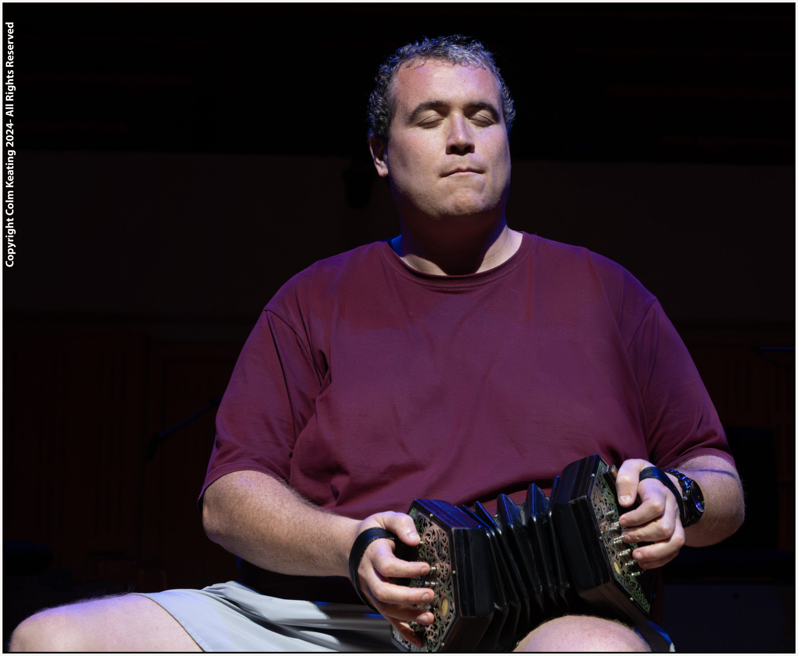 A man in a maroon shirt sits playing a concertina on his lap