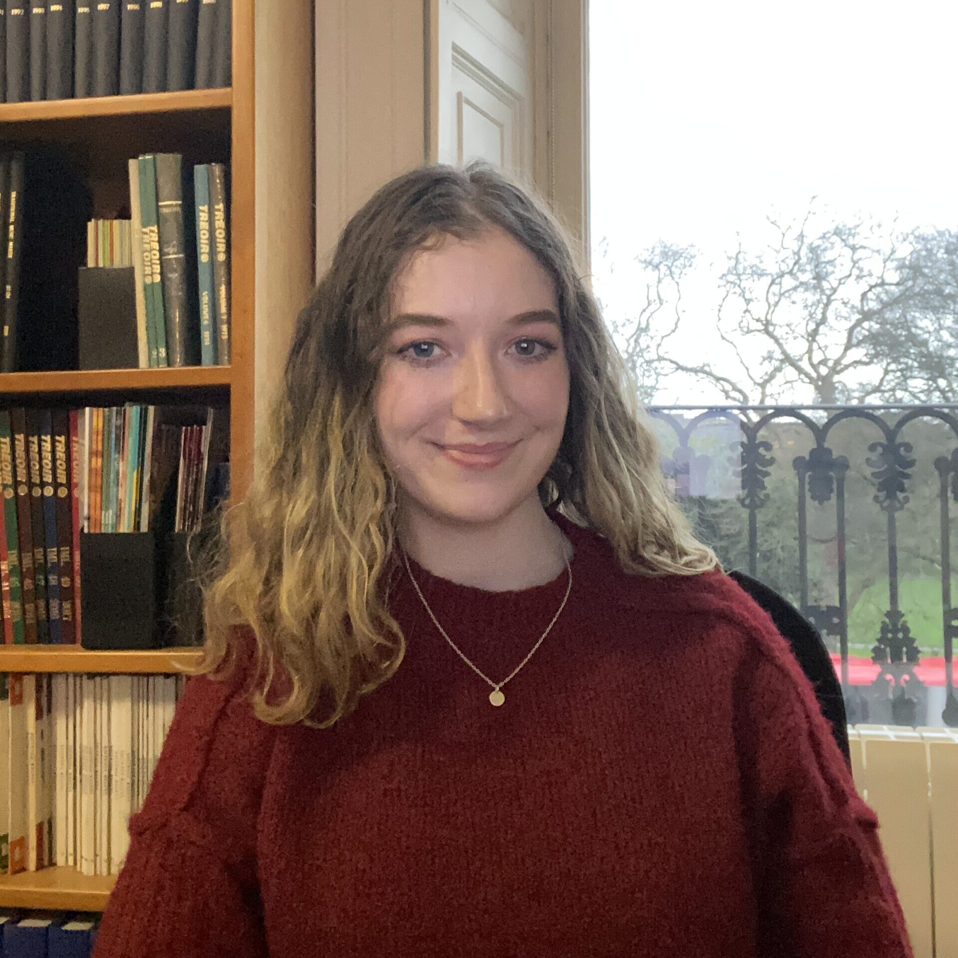 a woman wearing a red jumper sitting in front of a bookshelf and a window.