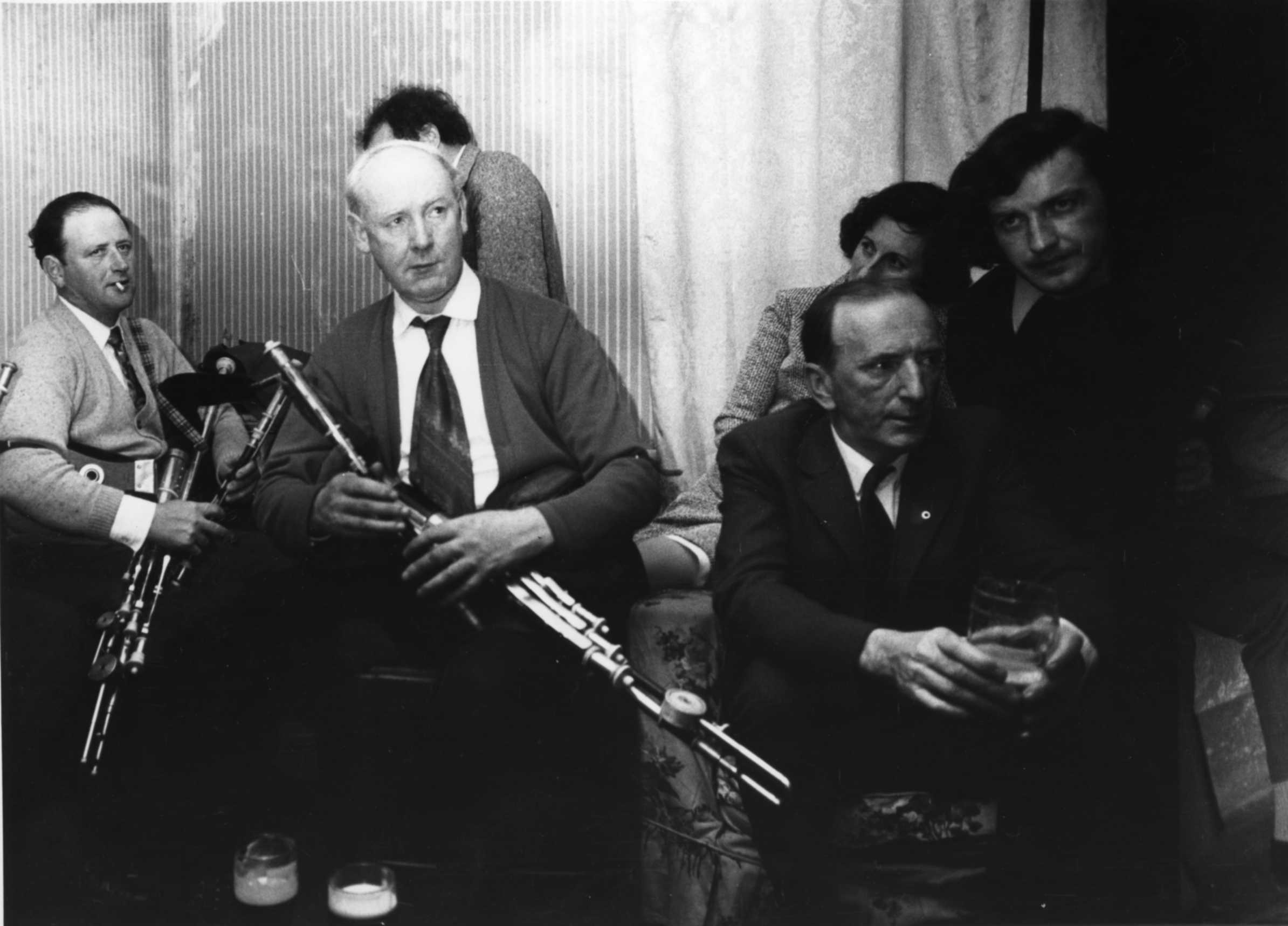 Sean Seery, seated playing pipes; Willie Clancy, seated playing pipes; Seán 'ac Dhonncha, seated holding drink; Unidentified woman (wife of Seán 'ac Dhonncha), seated behind Sean 'ac Dhonncha; Liam Óg Ó Floinn, seated. Photo by Mal Whyte, Cnuasach an Bhreathnaigh