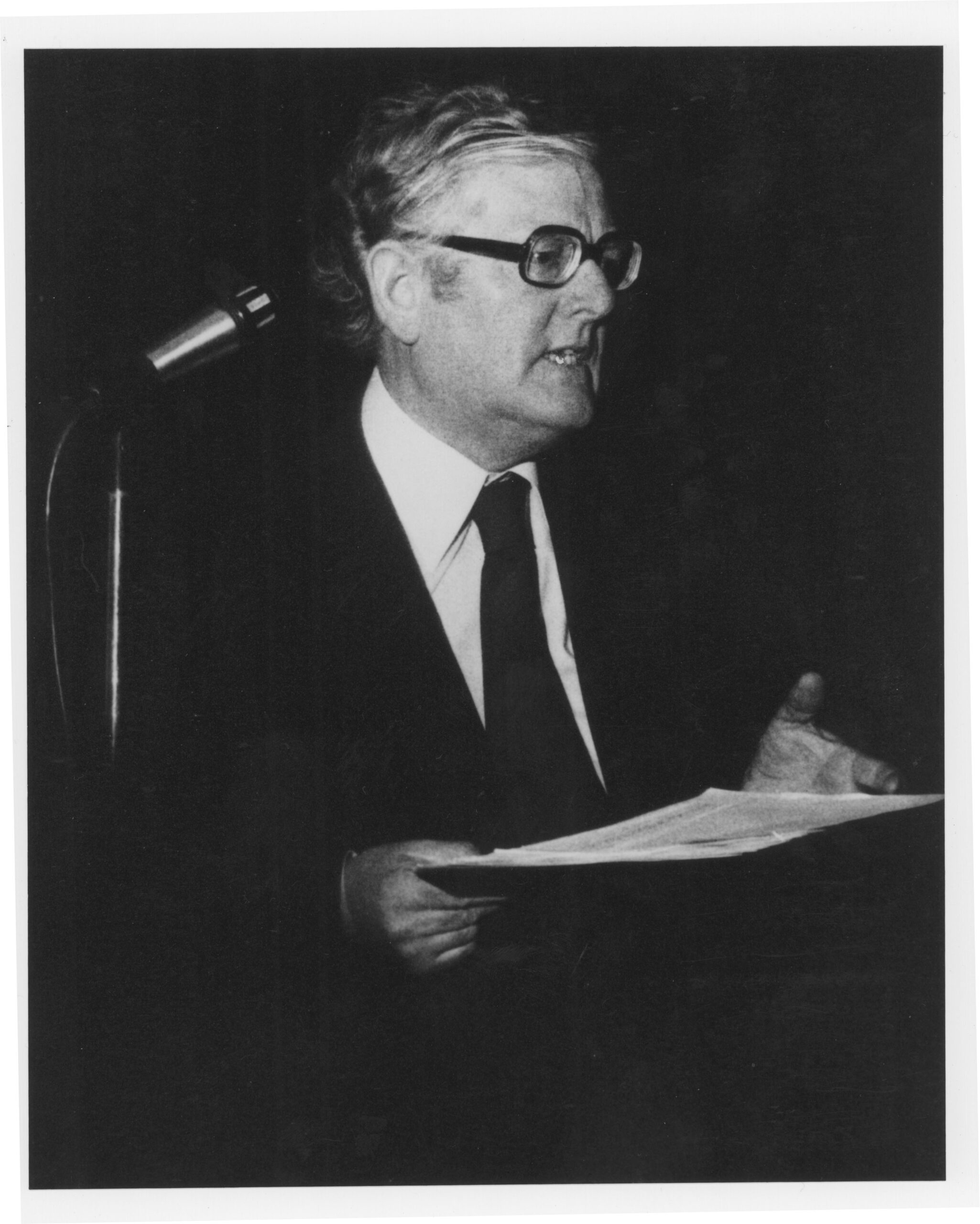 Black and white photo of Breandán Breathnach speaking in front of a microphone wearing a suit.
