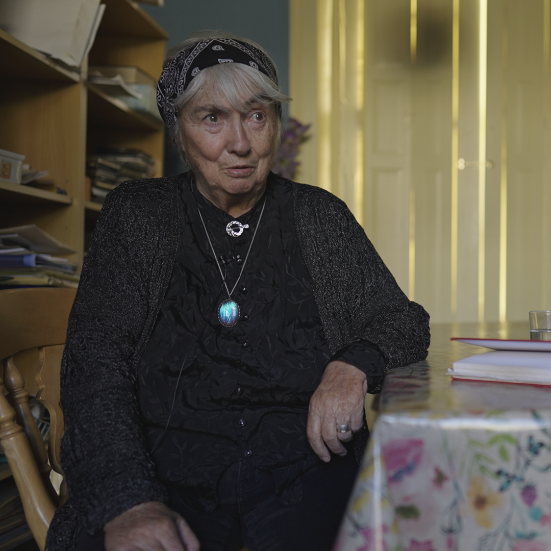 A woman with white hair sits at a table in a kitchen.