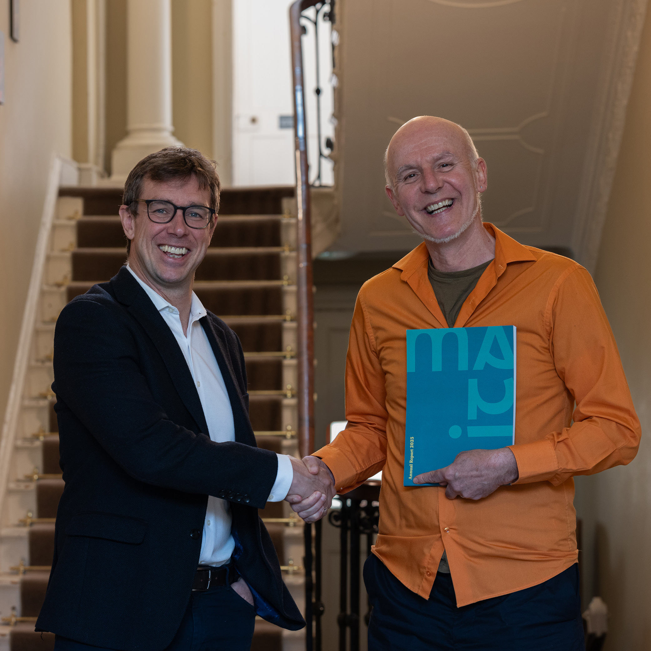 Two men stand in front of a stairwell shaking hands. The man on the right is holding a booklet that says ITMA on it.