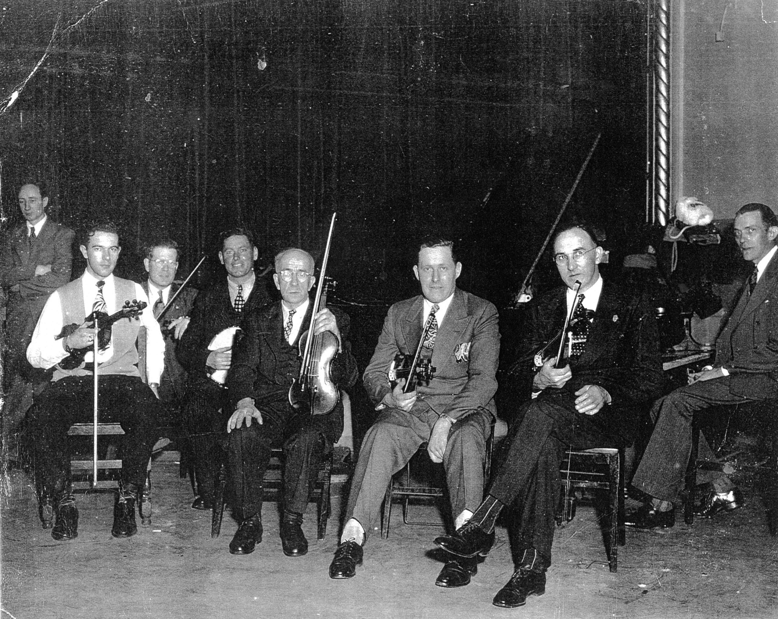 A black and white photo of a group of men in suits sitting holding fiddles on their laps.