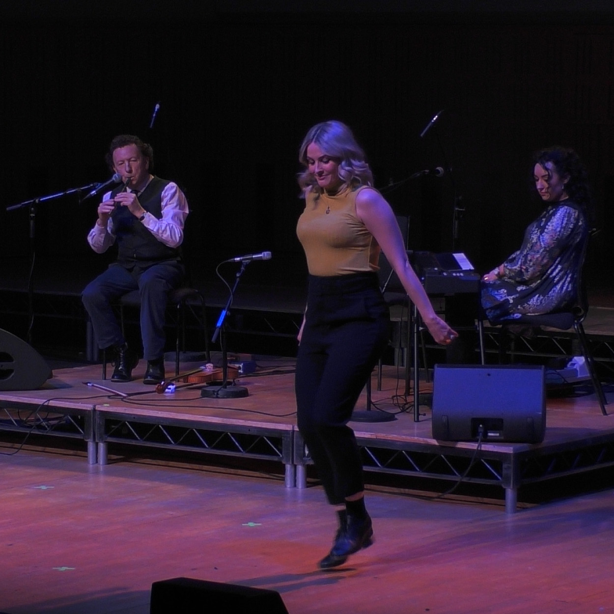 A woman dances on a stage, in the background behind her is a man playing the tin whistle and a woman sitting at a piano.
