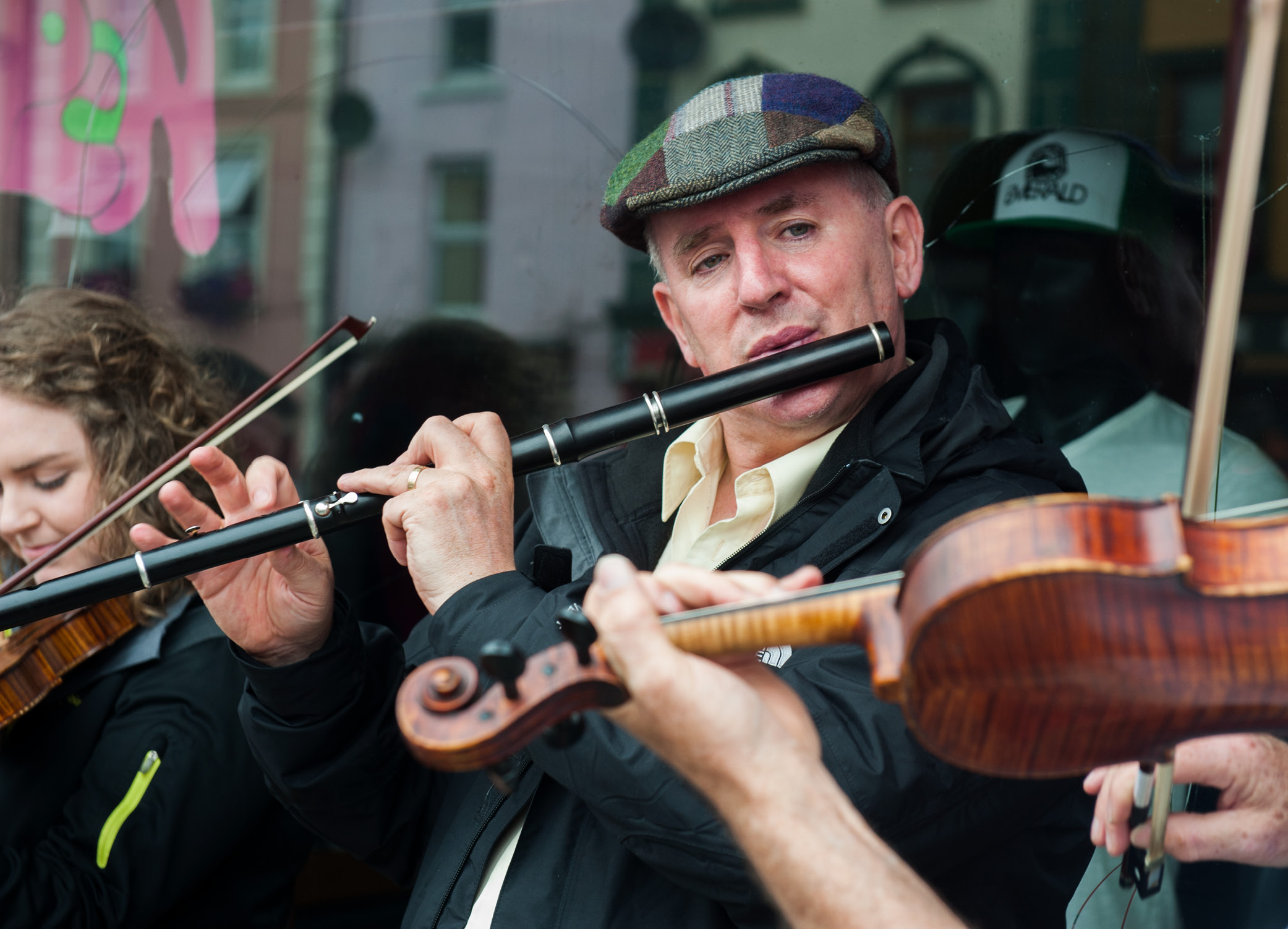 A man in a flat cap plays a wooden flute, with a person playing the fiddle beside him and in front of him.