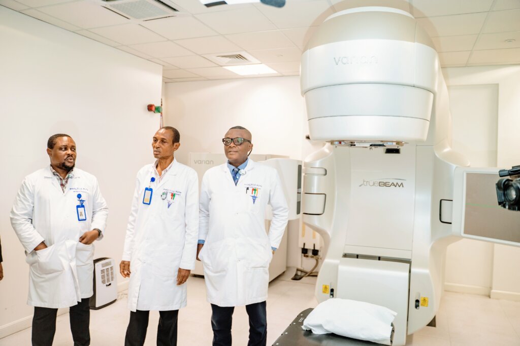 Three doctors in Marcelle Ruth lab coats stand beside a state-of-the-art Varian TrueBeam radiation therapy machine. The modern, clinical setting highlights Marcelle Ruth's commitment to providing advanced cancer treatment and exceptional healthcare services. The doctors' professional demeanor underscores the expertise and dedication of the Marcelle Ruth medical team.
