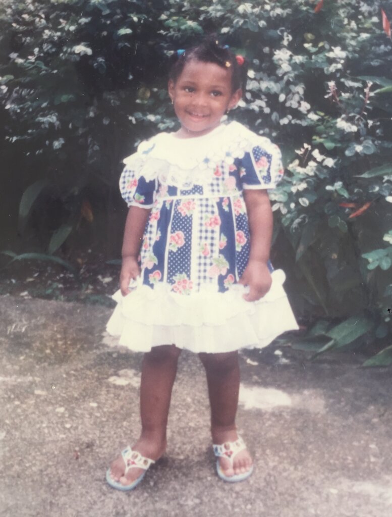 Me, wearing a blue dress with strawberry motifs, against a bush of flowers, in my childhood home in Port Harcourt, sometime in 2001/2002.