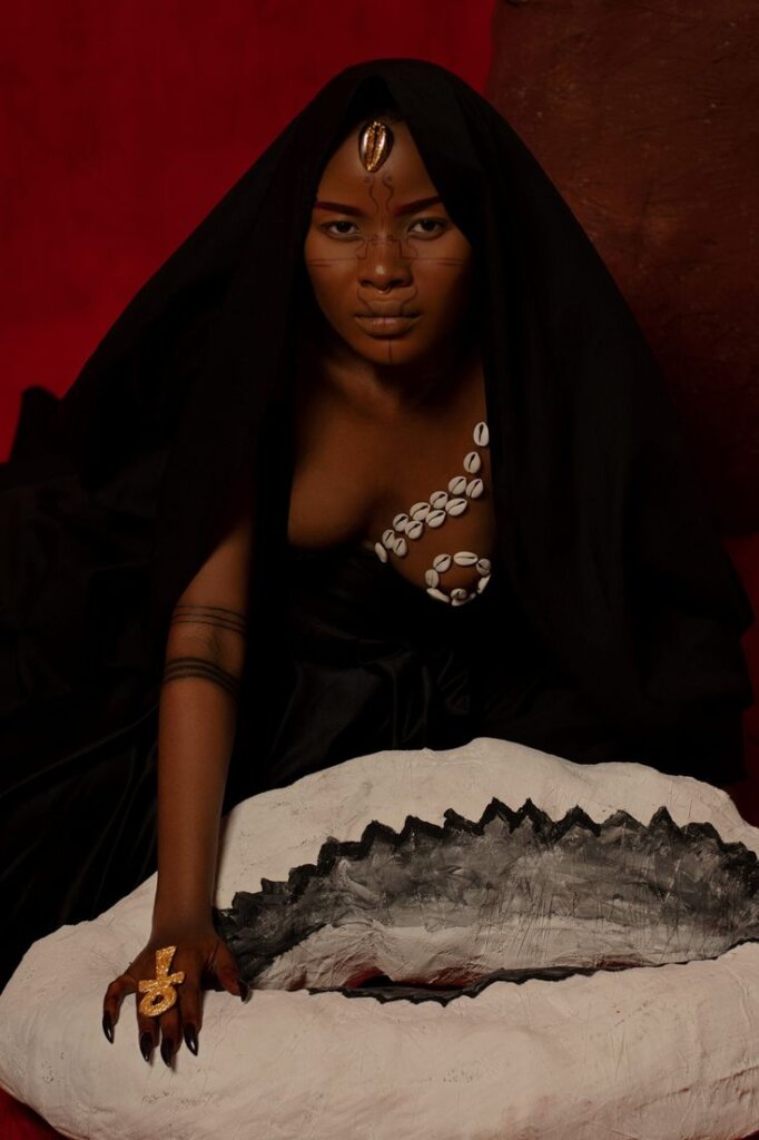 Black women wearing a head dress and cowrie body detailing against a red background via Pinterest
