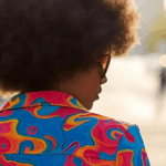 Style anxiety; close up shot of the back view of a woman with an afro, wearing a colourful jacket by Ampli Ficador via Unsplash