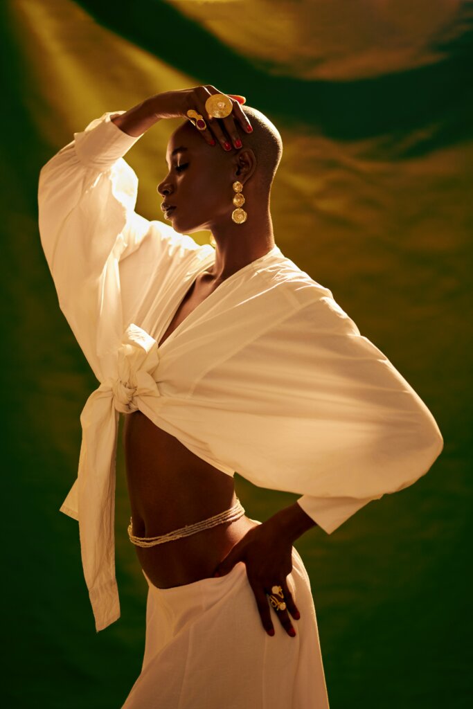 Black woman as divine; A Black woman with a shaved head, striking a fierce pose. She is wearing linen and gold jewellery.