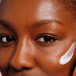 A dark skinned woman with a moisturiser swatch on her right cheek