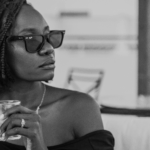 Black and white image of a Black woman wearing dark sunglasses and holding a glass by Lisa Marie-Theck via Unsplash
