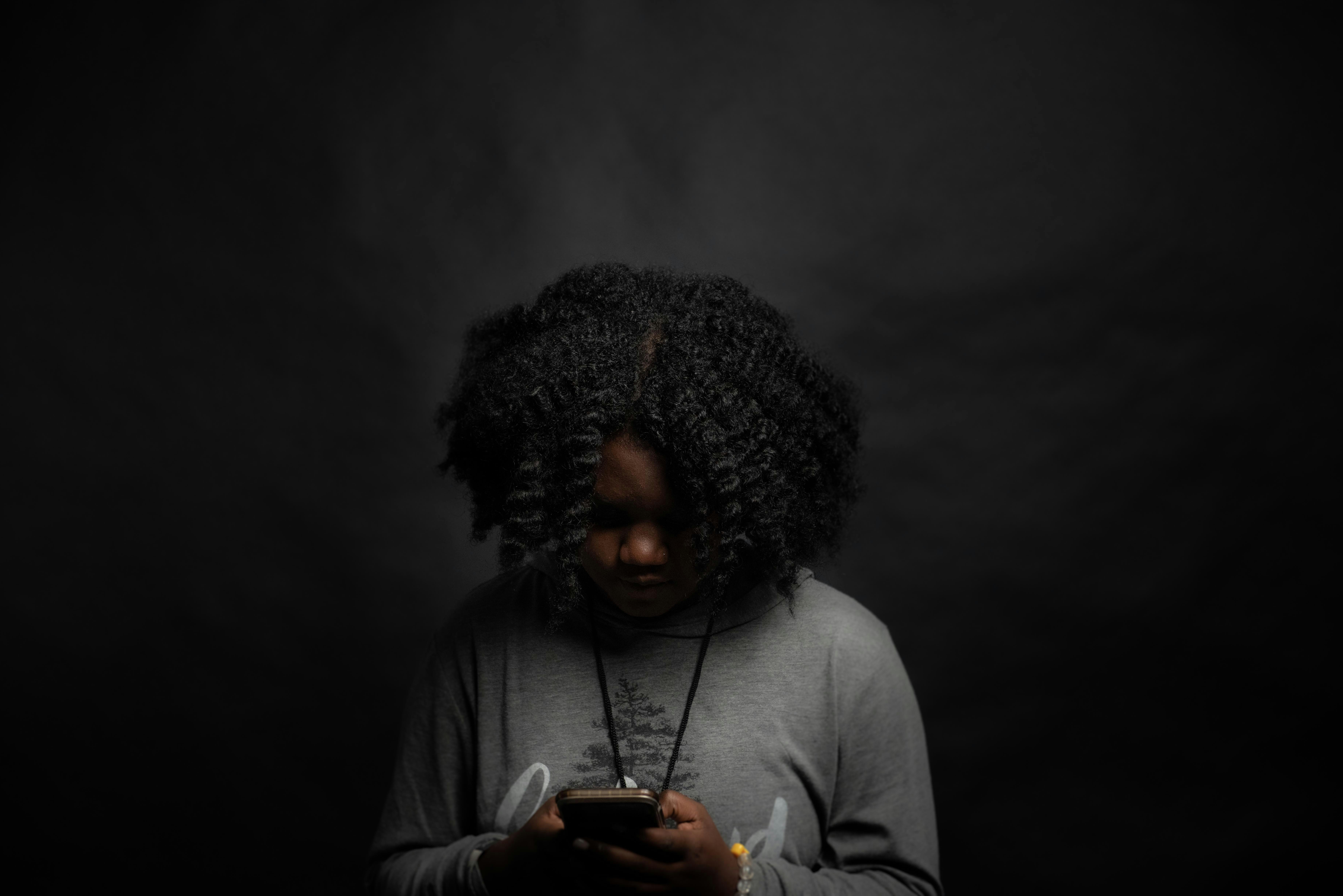 A dark image of a black woman looking sadly down at her phone by Chad Madden via Unsplash