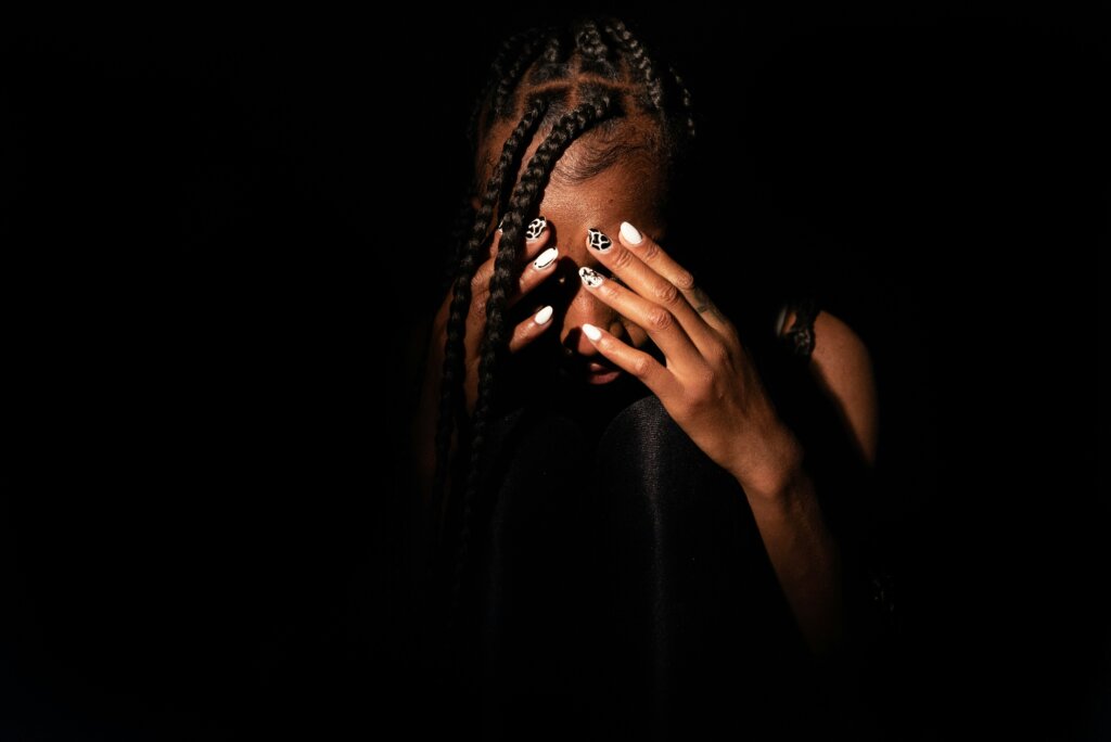 A Black woman with braids in a dark room. She has her hands placed over her eyes, and she looks forlorn.