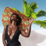 A black woman wearing a large kente beach hat with palm trees behind her by William Boateng via Unsplash