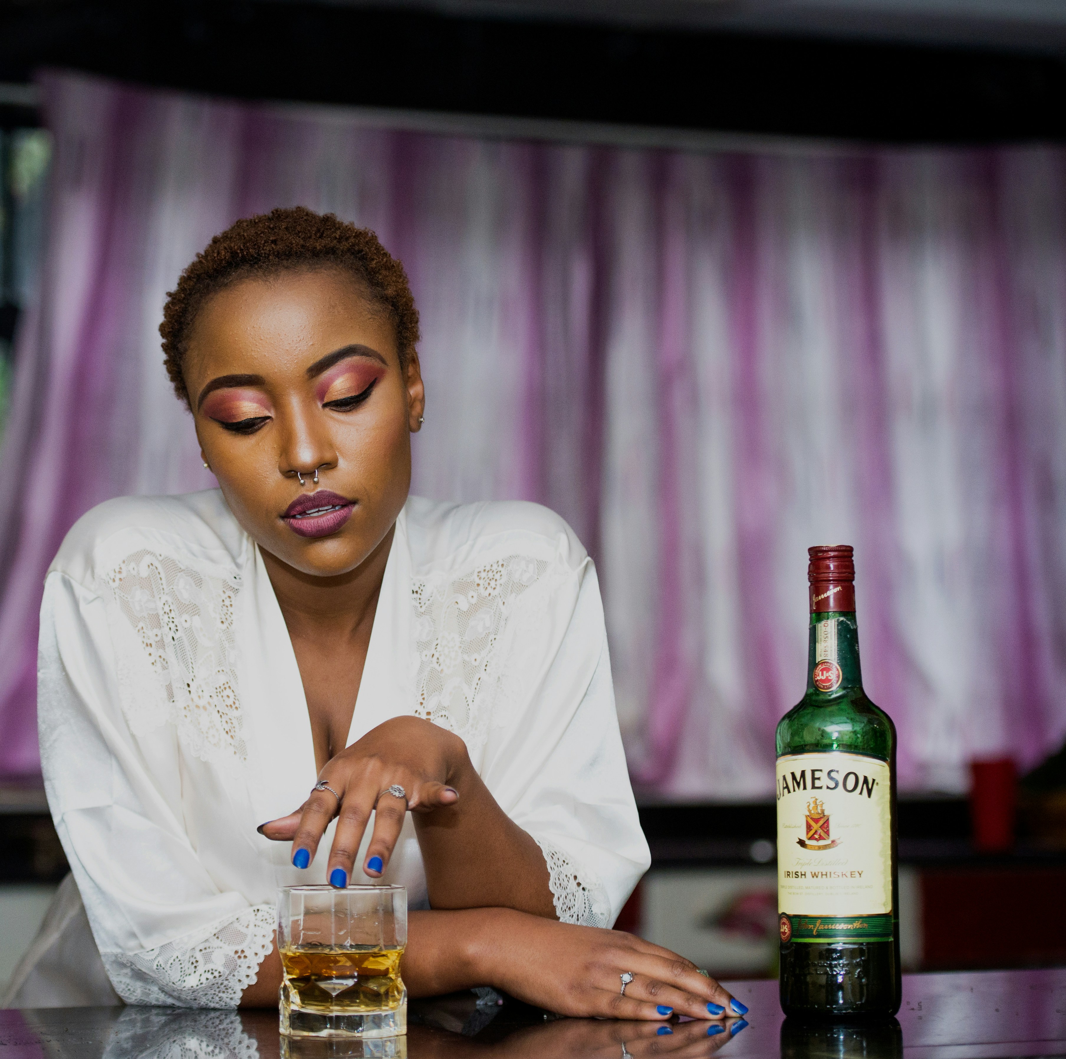 Black woman leaning against a table with a lowball half filled with whiskey in front of her. Her fingers her on the rim, and she's looking into the glass, a bottle of Jameson Whiskey is on the table beside her by Terricks Noah via Unsplash