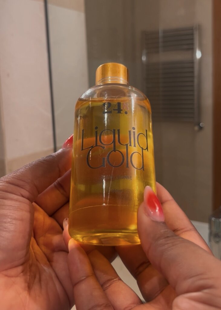 Two hands holding “Liquid Gold” Body Oil by 24.8 Skin by Aisha against a bathroom mirror