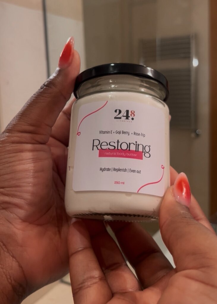Two hands holding “Restoring” Body Scrub by 24.8 Skin by Aisha against a bathroom mirror