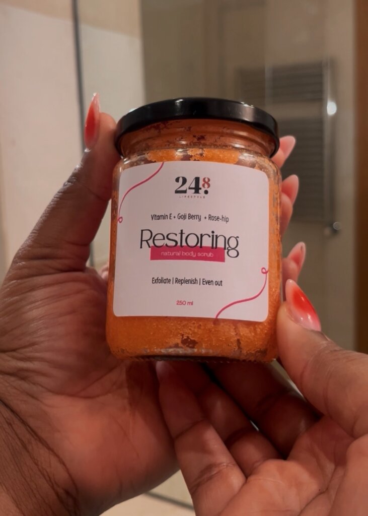 Two hands holding “Restoring” Body Scrub by 24.8 Skin by Aisha against a bathroom mirror