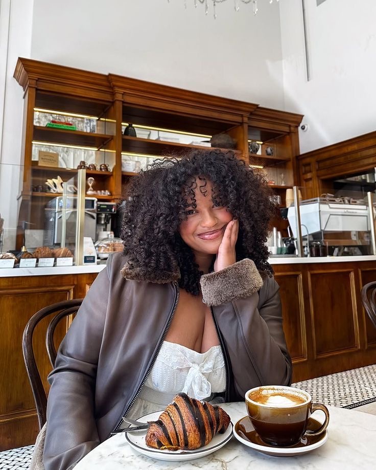 Black woman sitting at a table in a café with a croissant and coffee via Zee on Pinterest