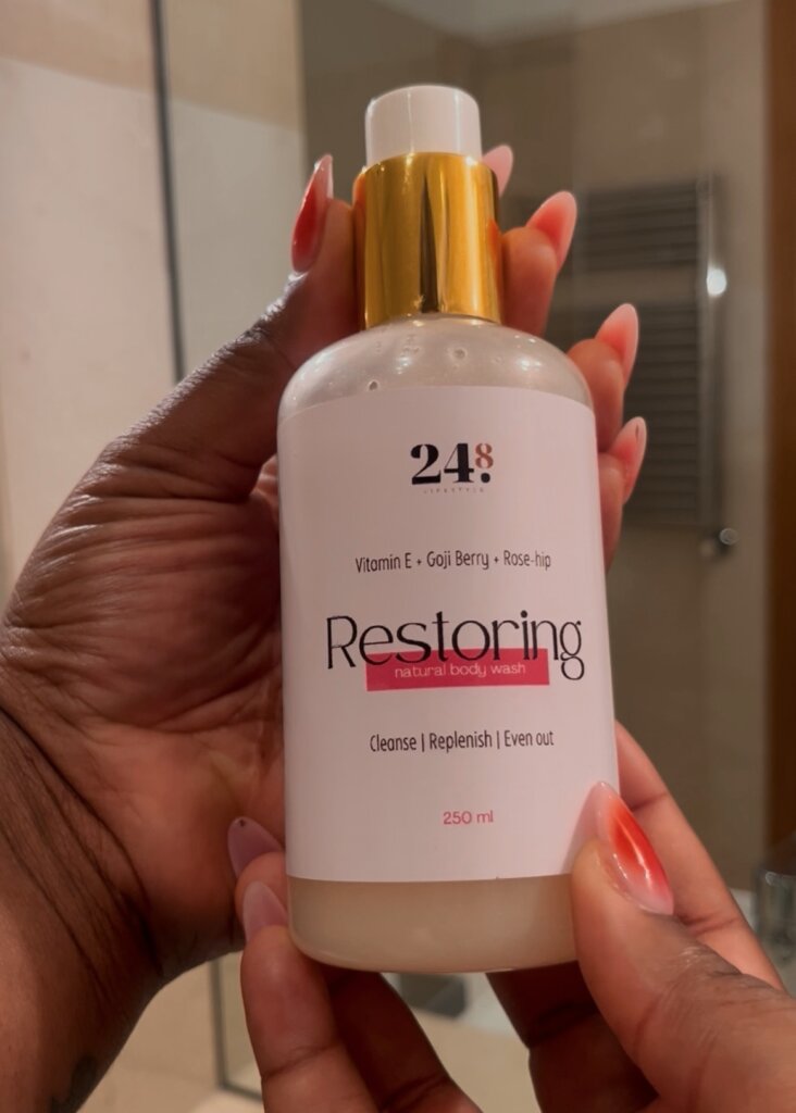 Two hands holding “Restoring” Body Wash by 24.8 Skin by Aisha against a bathroom mirror