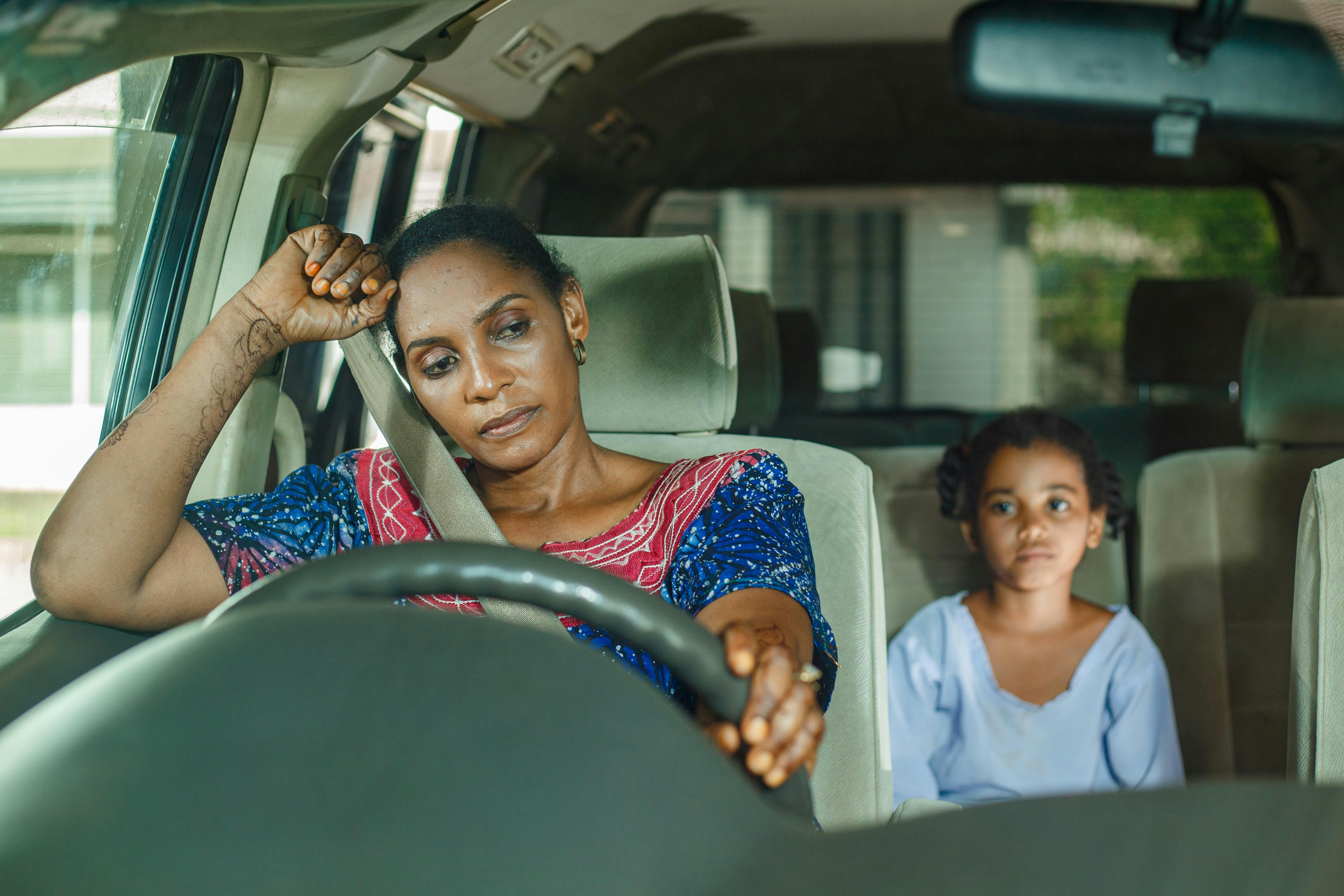 Black woman with a tired expression driving a car with a little girl in the backseat by Ali Mkumbwa via Unsplash
