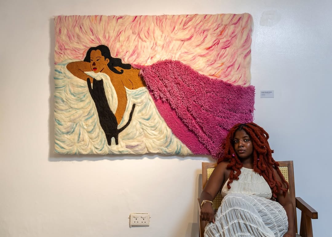 Cynthia Ugwudike seated beside her artwork at her show “In the eyes of my lover” via @cynugwu via Instagram