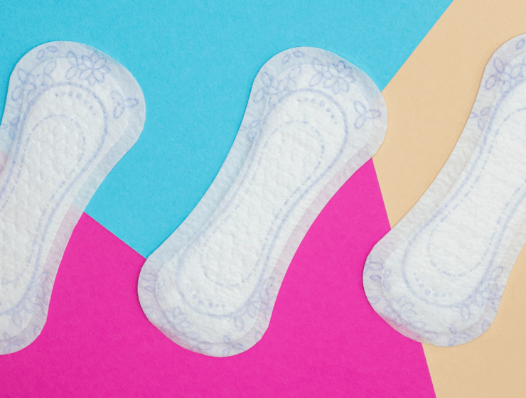 Three white sanitary pads with subtle floral patterns displayed on a bold geometric background in blue, pink, and beige.