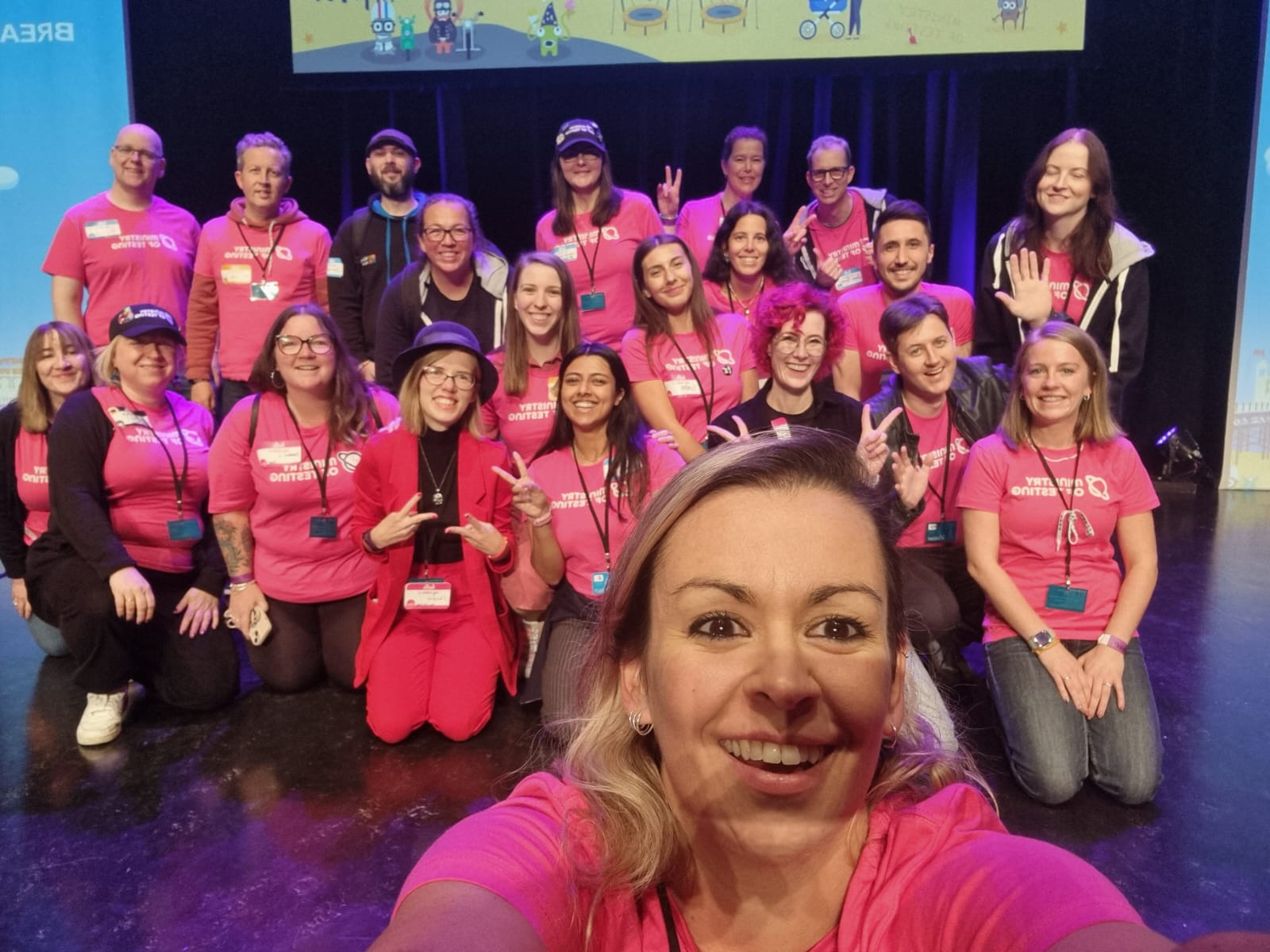 A group of several people kneeling and standing up on a conference stage. There is a woman at the front taking a selfie. Most people wear pink t-shirts with a Ministry of Testing logo. Most people are smiling.  
