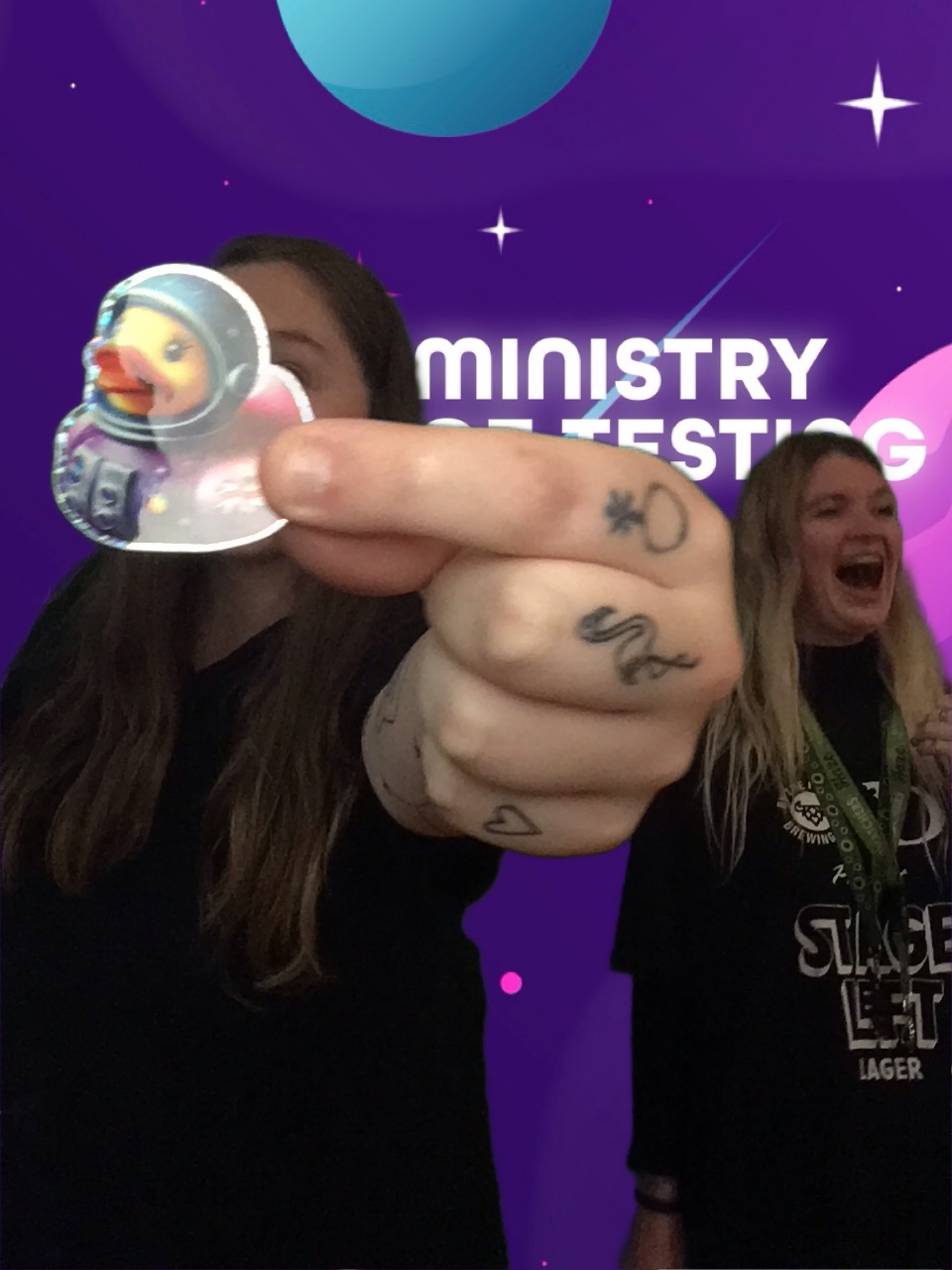 Now this picture has more of a messy feel but compositionally it pops! In the foreground a hand shows the spaceduck shiny sticker to the camera whilst 2 TestBash attendees laugh uncontrollably. The light isn't great and it's obviously a bit of a mistake but it really encapsulates the joy and lightness of the MoT community. 

Background: The green screen backdrop depicts a simple illustrated outer space scene. The main colour is purple, with cute stars and planets in shades of pink, blue, and white. About a third of the way down the screen, a logo appears. It reads "Ministry of Testing" in bold white text with a blue shooting star behind it. Next to the text, in the same bold white style, is a line drawing of the planet Saturn.
